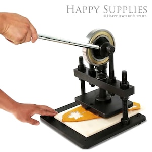May include: A black metal leather craft press with a hand crank and a white cutting mat. A yellow leather piece with a leaf cutout is on the mat.