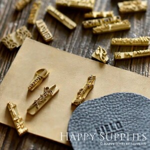 K&ouml;nnte beinhalten: Eine Sammlung von goldfarbenen Metallstempeln mit verschiedenen Designs und Beschriftungen, darunter "Hello" und "Happy Jewelry Supplies". Die Stempel sind auf einer braunen Lederfl&auml;che angeordnet.