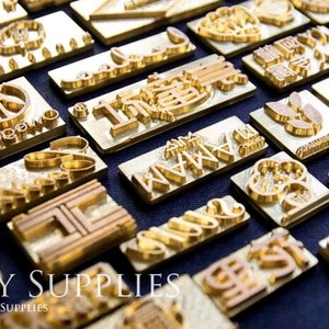 K&ouml;nnte beinhalten: Eine Sammlung von goldfarbenen Metallstempeln mit verschiedenen Designs, darunter Symbole, Buchstaben und W&ouml;rter. Die Stempel sind auf einer blauen Oberfl&auml;che angeordnet. HAPPY SUPPLIES &copy; HAPPY JEWELRY SUPPLIES