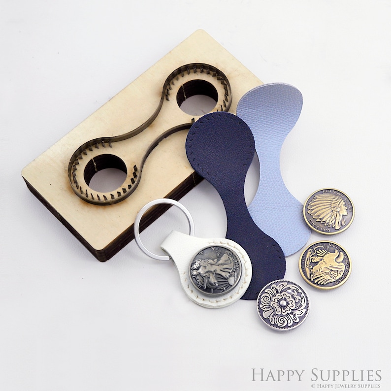 May include: A wooden punch tool with two circular cutouts, used for crafting leather keychains. The image also shows a white leather keychain with a silver coin design, two blue leather pieces, and three metal buttons with different designs.