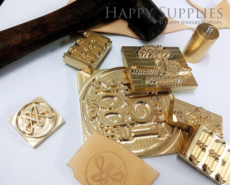 K&ouml;nnte beinhalten: Ein Satz Stempel aus Messing f&uuml;r die Lederverarbeitung. Die Stempel zeigen verschiedene Designs, darunter Zahlen, Buchstaben und dekorative Muster. Die Stempel sind auf einer hellbraunen Lederoberfl&auml;che angeordnet, zusammen mit einem Holzhammer und einem Messinggriff.