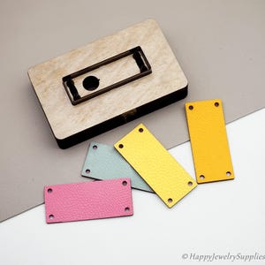 May include: A wooden die-cutting tool with a metal frame, alongside several rectangular leather pieces in pink, blue, and yellow. Each leather piece has four pre-punched holes, suggesting a crafting or jewelry-making theme.