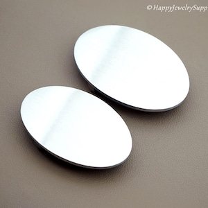 May include: Two oval-shaped, silver-colored metal jewelry components. The components have a brushed metal finish and are set against a neutral brown background. The image is well-lit, highlighting the reflective surfaces.