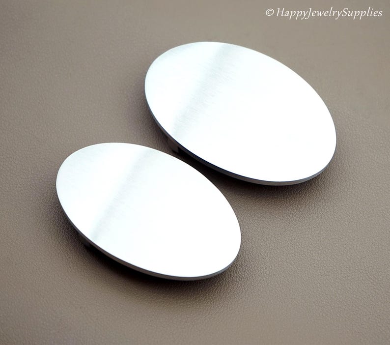 May include: Two oval-shaped, silver-colored metal jewelry components. The components have a brushed metal finish and are set against a neutral brown background. The image is well-lit, highlighting the reflective surfaces.