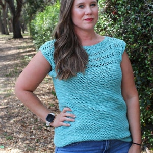 May include: A turquoise crochet top with a textured design. The top has a round neckline and cap sleeves. The image is taken outdoors with a natural background of trees and foliage.