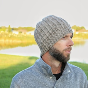 May include: A gray knitted beanie with a ribbed texture. The beanie is worn by a person with a beard and a gray sweater.