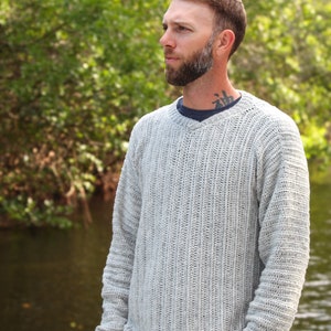 May include: A man wearing a light gray knitted sweater with a v-neck and long sleeves. He is standing outdoors with a body of water in the background.
