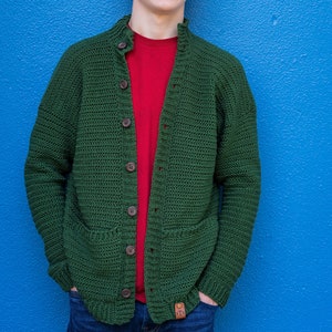 May include: A green crocheted cardigan with a red t-shirt underneath. The cardigan has a button closure and two pockets.
