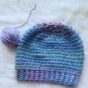 Puede incluir: Un gorro de punto de ganchillo en tonos azules, morados y rosas. El gorro está hecho con un punto texturizado y tiene un diseño simple y clásico.