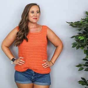 May include: A woman wearing a coral crochet tank top with a scoop neck and a pair of blue denim shorts.
