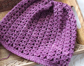 Può includere: Un berretto lavorato a maglia di colore viola con un motivo traforato. Il berretto è appoggiato su un vassoio di legno con un design intagliato.