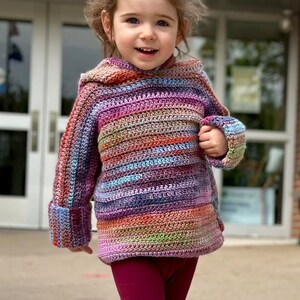 May include: A young child wearing a colorful striped crocheted sweater with a hood and maroon leggings.