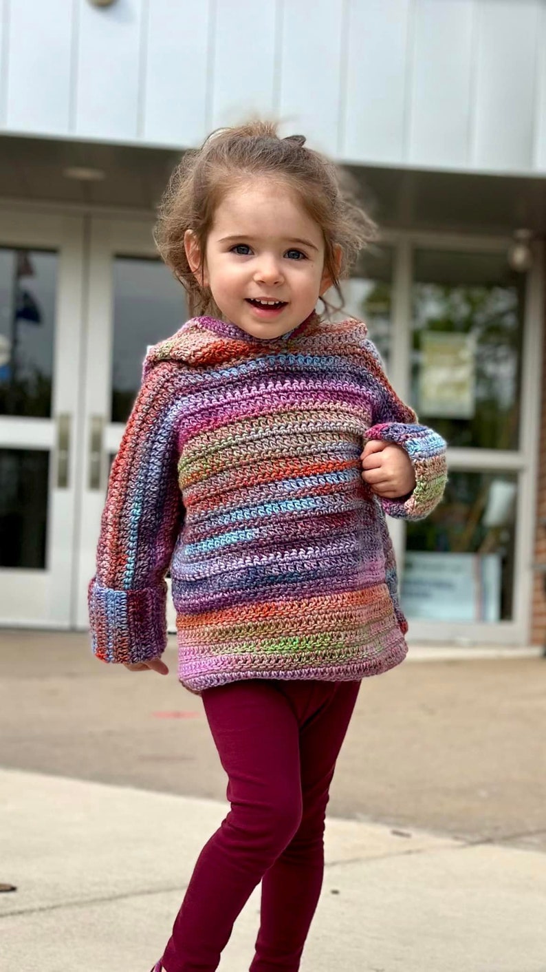 May include: A young child wearing a colorful striped crocheted sweater with a hood and maroon leggings.