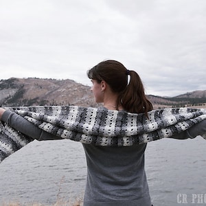 May include: A gray, white, and black crocheted shawl is held open against a backdrop of a lake and mountains. The shawl features a striped pattern with openwork sections. The person is wearing a gray long-sleeved shirt.
