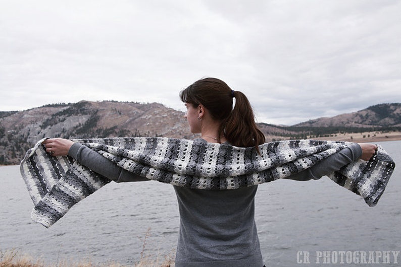 May include: A gray, white, and black crocheted shawl is held open against a backdrop of a lake and mountains. The shawl features a striped pattern with openwork sections. The person is wearing a gray long-sleeved shirt.