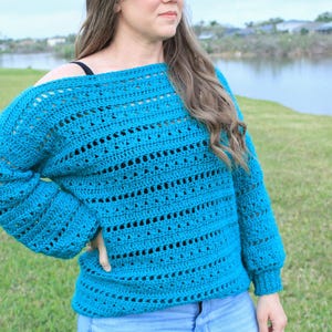 Puede incluir: Un suéter de crochet azul turquesa de corte holgado y mangas largas. El suéter tiene un delicado patrón de punto abierto.