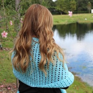 May include: A light blue, open-weave crochet shawl. The shawl is draped over the shoulders. The person is facing away from the camera, with long, wavy, light brown hair. The background includes a lake, trees, and pink flowers.
