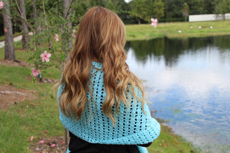 May include: A light blue, open-weave crochet shawl. The shawl is draped over the shoulders. The person is facing away from the camera, with long, wavy, light brown hair. The background includes a lake, trees, and pink flowers.
