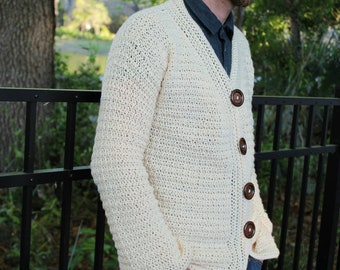 May include: A cream-colored crocheted cardigan with brown buttons. The cardigan is worn by a person standing in front of a black metal fence.