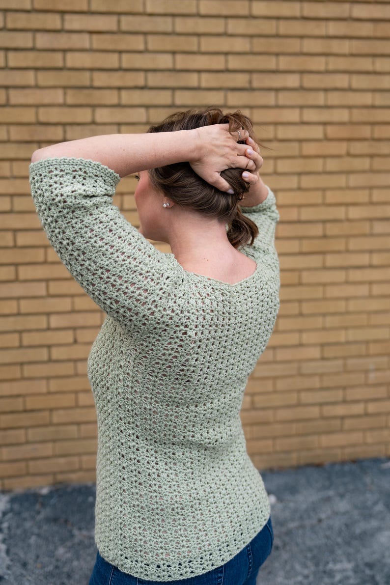 May include: A light green crochet top with long sleeves and a scoop neck. The top is made of a loosely woven material, and the person is wearing blue jeans. The background is a brick wall.