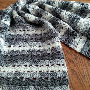 May include: A handmade crochet blanket in shades of gray and white. The blanket features a textured pattern with alternating stripes of solid and openwork stitches. The soft, cozy blanket is draped on a wooden surface.