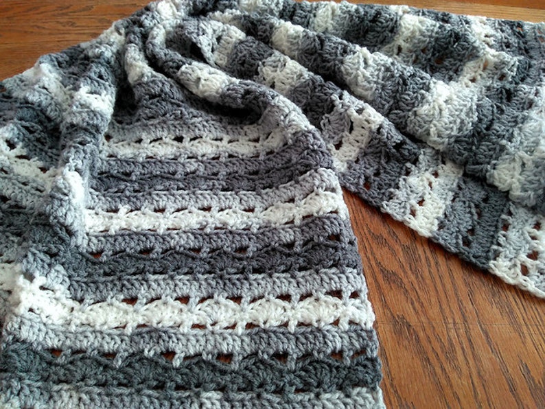 May include: A handmade crochet blanket in shades of gray and white. The blanket features a textured pattern with alternating stripes of solid and openwork stitches. The soft, cozy blanket is draped on a wooden surface.