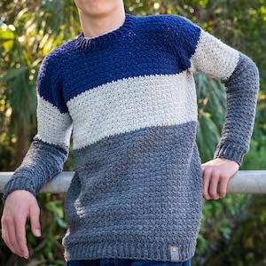 Könnte beinhalten: Ein farbblockierter Pullover in Blau-, Grau- und Weißtönen. Der Pullover hat einen Rundhalsausschnitt und lange Ärmel. Der untere Teil des Pullovers hat ein kleines Etikett mit den Initialen "BTB".
