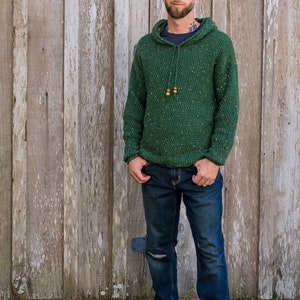 May include: A man wearing a green speckled hooded sweater with brown wooden beads on the drawstring. He is wearing blue jeans and gray high-top sneakers.