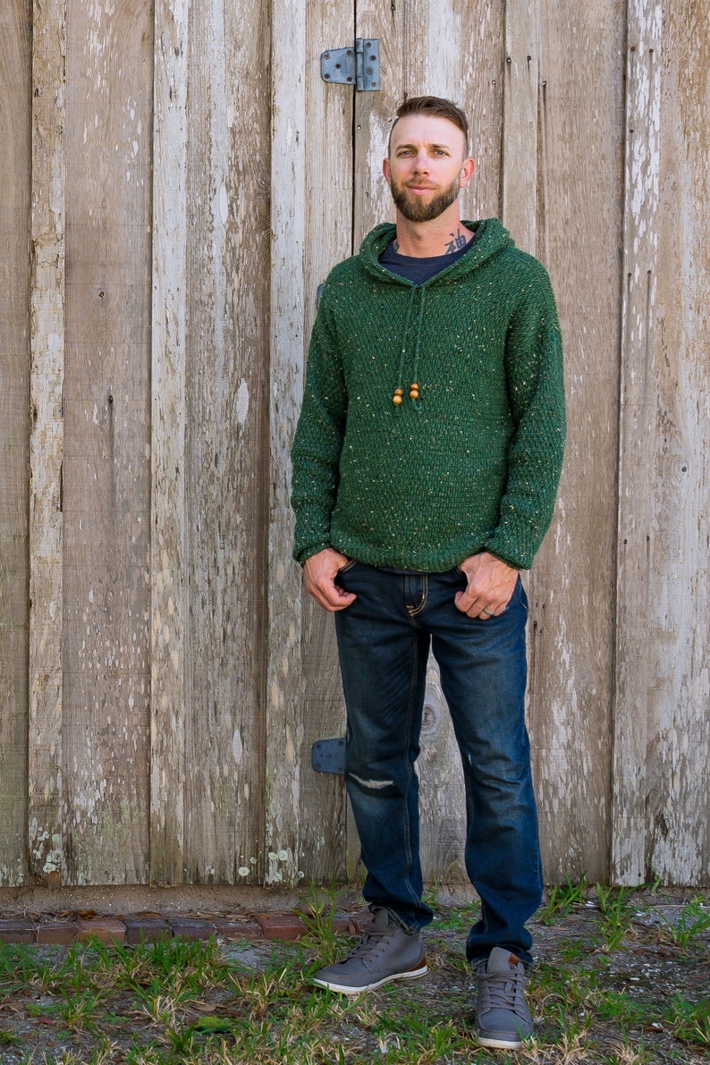 May include: A man wearing a green speckled hooded sweater with brown wooden beads on the drawstring. He is wearing blue jeans and gray high-top sneakers.