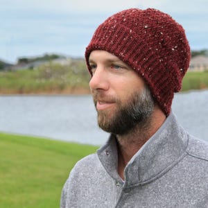 Puede incluir: Un hombre con una boina de punto roja con un patrón moteado. Lleva un suéter gris con cuello de botón.