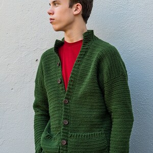 May include: A green crocheted cardigan with a button closure and a pocket on the front. The cardigan is worn over a red t-shirt and blue jeans.