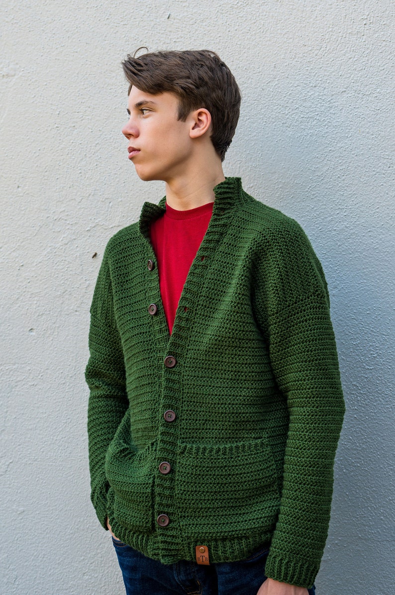 May include: A green crocheted cardigan with a button closure and a pocket on the front. The cardigan is worn over a red t-shirt and blue jeans.
