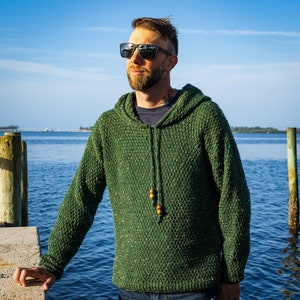 May include: A man wearing a green hooded sweater with a speckled texture. He is standing outdoors near a body of water and wearing sunglasses.