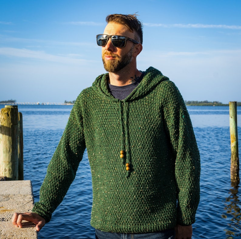 May include: A man wearing a green hooded sweater with a speckled texture. He is standing outdoors near a body of water and wearing sunglasses.