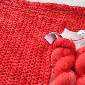 May include: A close-up of a red crocheted fabric with two skeins of red yarn. The yarn has white labels that read "AUDINE WOOLS". The crocheted fabric is a solid red color.