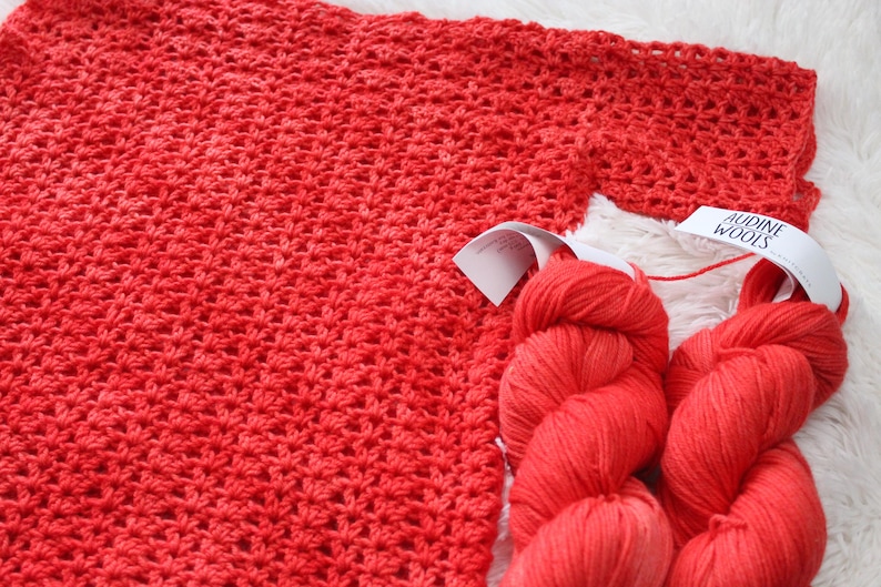 May include: A close-up of a red crocheted fabric with two skeins of red yarn. The yarn has white labels that read "AUDINE WOOLS". The crocheted fabric is a solid red color.