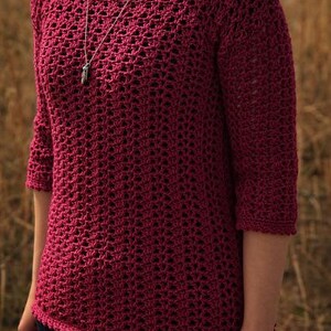 May include: A burgundy crochet top with three-quarter sleeves. The open weave design of the top is visible. A silver necklace with a pendant is worn. The top is paired with dark blue jeans.