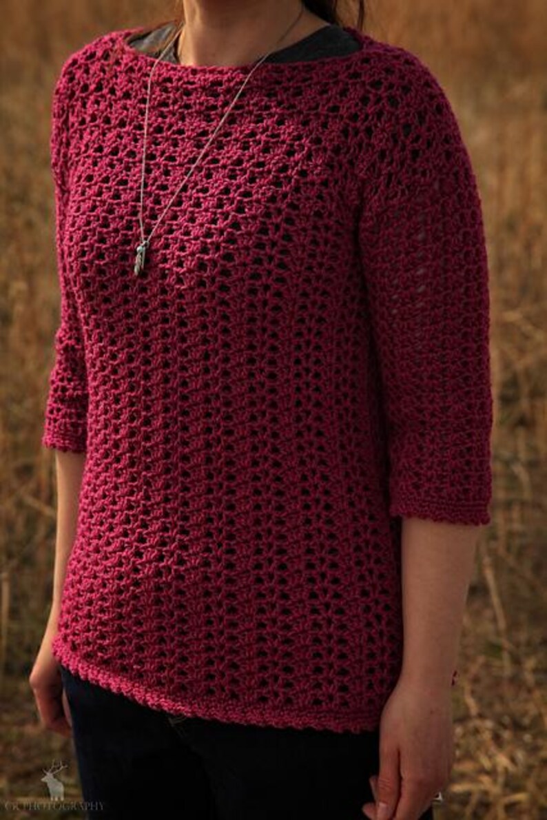May include: A burgundy crochet top with three-quarter sleeves. The open weave design of the top is visible. A silver necklace with a pendant is worn. The top is paired with dark blue jeans.