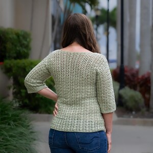 May include: A light green, crocheted top with three-quarter sleeves. The top has a textured, open weave design and a fitted silhouette. The person is wearing blue jeans. The image is taken outdoors.