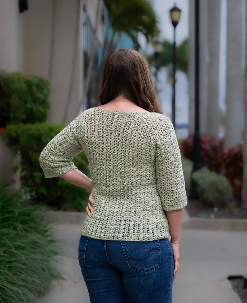 May include: A light green, crocheted top with three-quarter sleeves. The top has a textured, open weave design and a fitted silhouette. The person is wearing blue jeans. The image is taken outdoors.