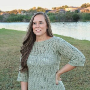 May include: A light green crochet sweater with three-quarter sleeves and a boat neck. The sweater is paired with blue jeans. The image is taken outdoors with a lake and trees in the background.