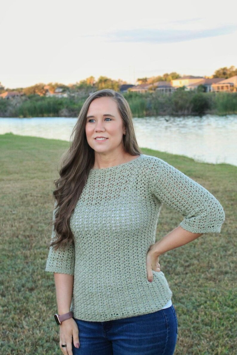 May include: A light green crochet sweater with three-quarter sleeves and a boat neck. The sweater is paired with blue jeans. The image is taken outdoors with a lake and trees in the background.