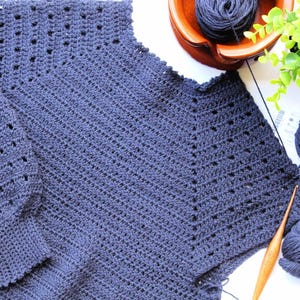 May include: A dark blue crocheted sweater with a loose fit and long sleeves. The sweater is made with a stitch pattern that creates a textured look. The sweater is laid flat on a white surface with yarn and a crochet hook.