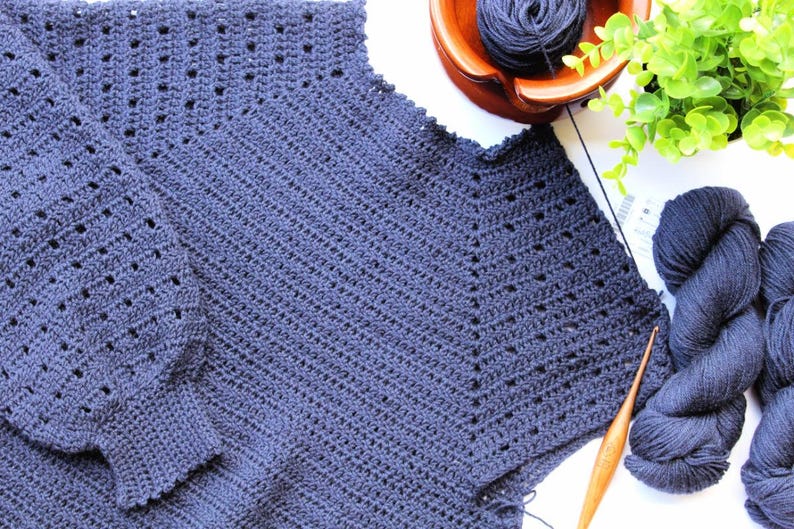 May include: A dark blue crocheted sweater with a loose fit and long sleeves. The sweater is made with a stitch pattern that creates a textured look. The sweater is laid flat on a white surface with yarn and a crochet hook.
