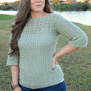 May include: A light green, crocheted top with three-quarter sleeves. The top is paired with blue jeans. The model is wearing a watch with a brown band. The background features a body of water and green grass.
