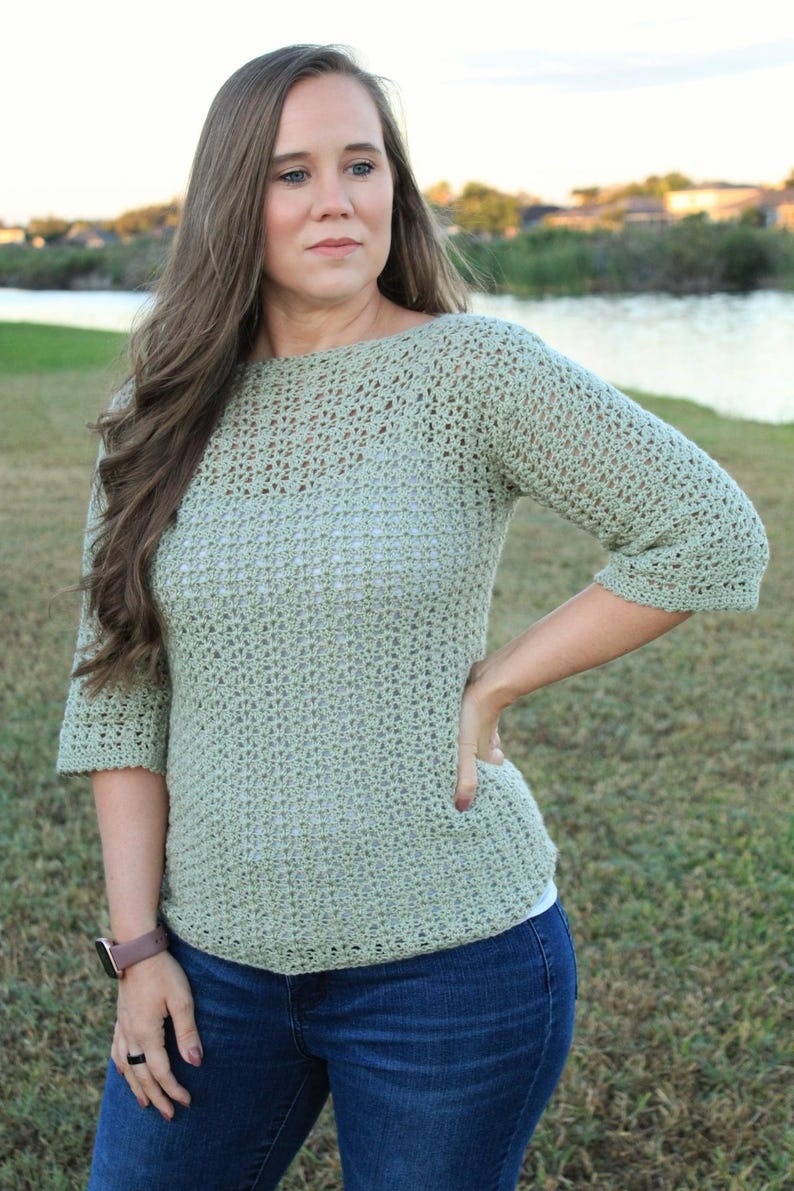 May include: A light green, crocheted top with three-quarter sleeves. The top is paired with blue jeans. The model is wearing a watch with a brown band. The background features a body of water and green grass.