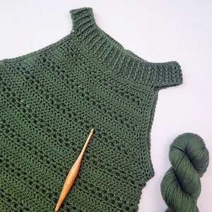 May include: A dark green, handmade crochet top with a textured stitch pattern and a wooden crochet hook. A skein of matching green yarn is also visible. The top has a ribbed neckline and shoulder straps, indicating a handcrafted item.
