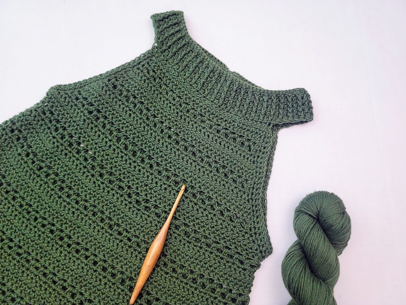 May include: A dark green, handmade crochet top with a textured stitch pattern and a wooden crochet hook. A skein of matching green yarn is also visible. The top has a ribbed neckline and shoulder straps, indicating a handcrafted item.