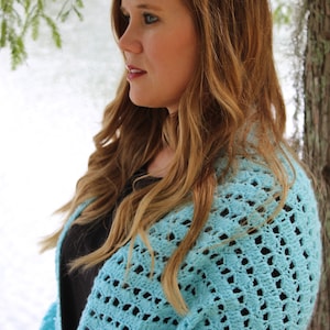 May include: A light blue, open-weave crochet shawl. The shawl is draped over the shoulders. The image shows a woman with long, wavy, light brown hair. The background includes a body of water and tree branches.