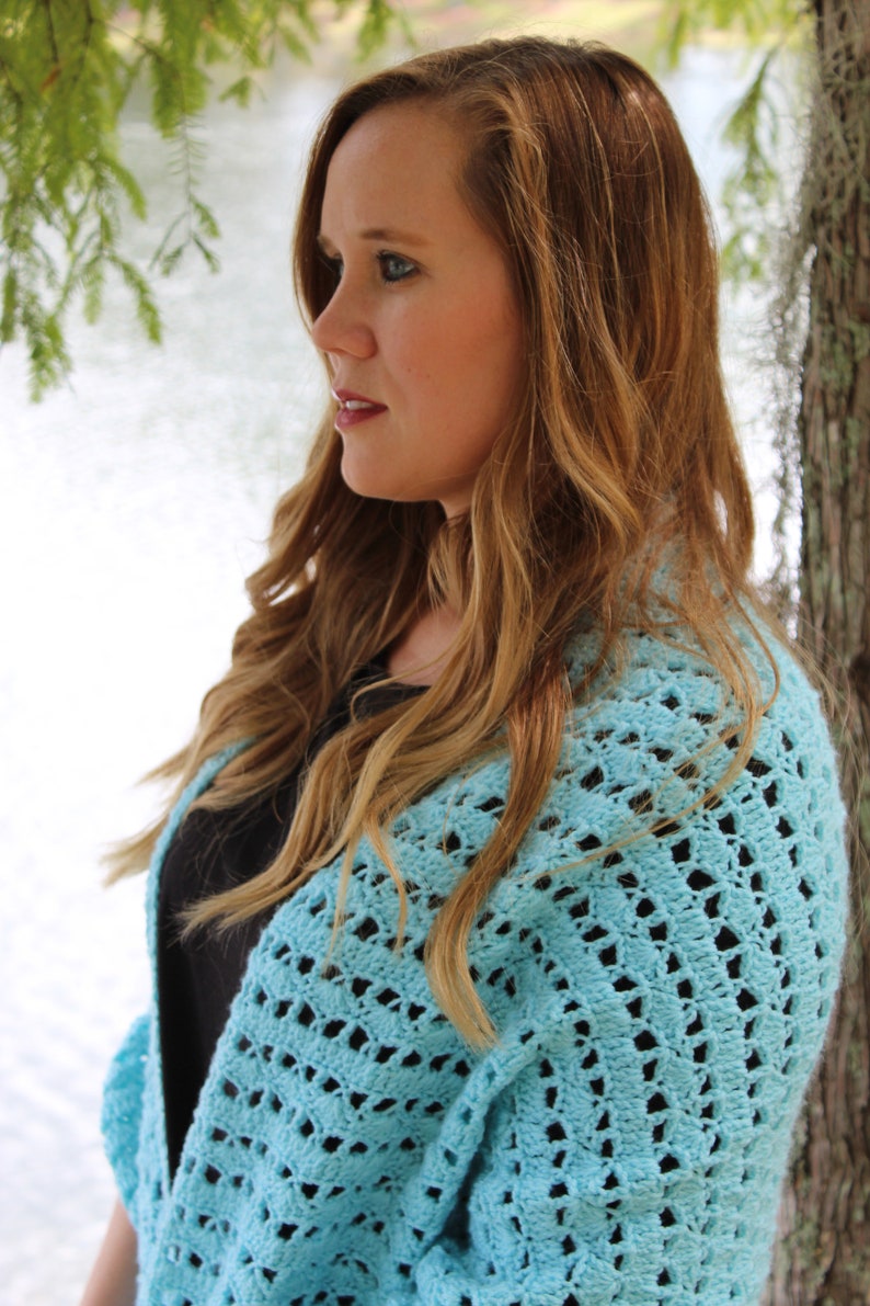 May include: A light blue, open-weave crochet shawl. The shawl is draped over the shoulders. The image shows a woman with long, wavy, light brown hair. The background includes a body of water and tree branches.
